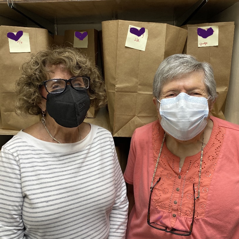 Two women stand in front of full paper grocery bags. ElderNet volunteers Rita and Rosemary pack bags for community members who are facing food insecurities. 